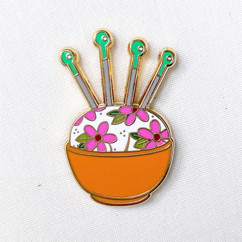 May include: Gold pin featuring a floral pincushion and four green and gold needles. The pincushion is in a small orange bowl.
