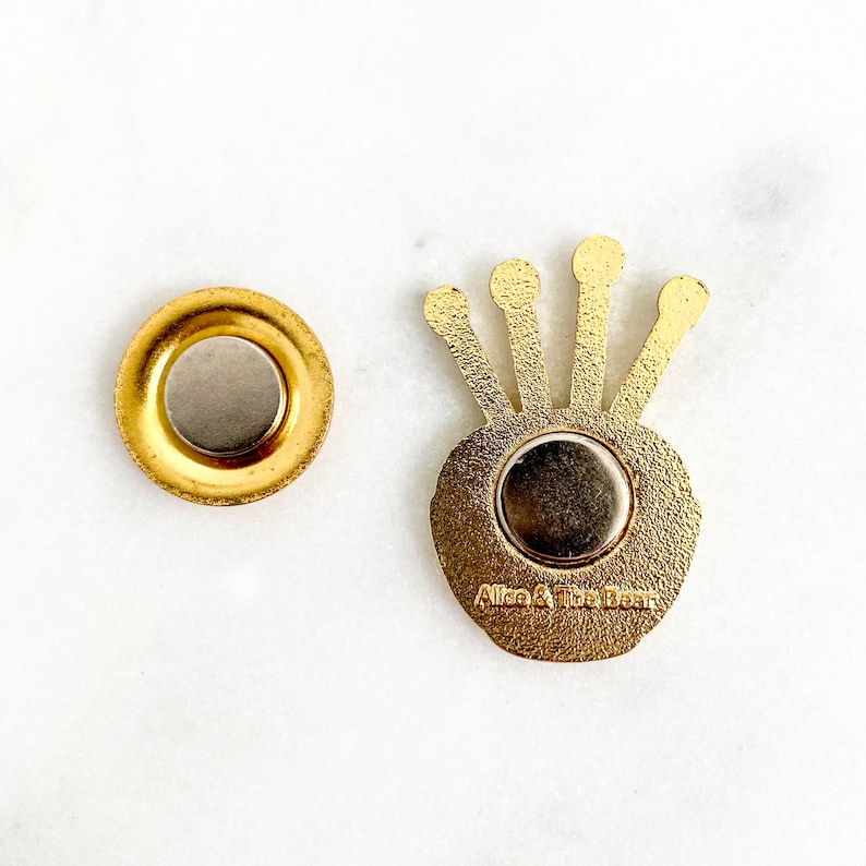 May include: Gold-coloured metal pin with a crown design and a magnetic back. The pin says "Alice & The Bear" on the back.