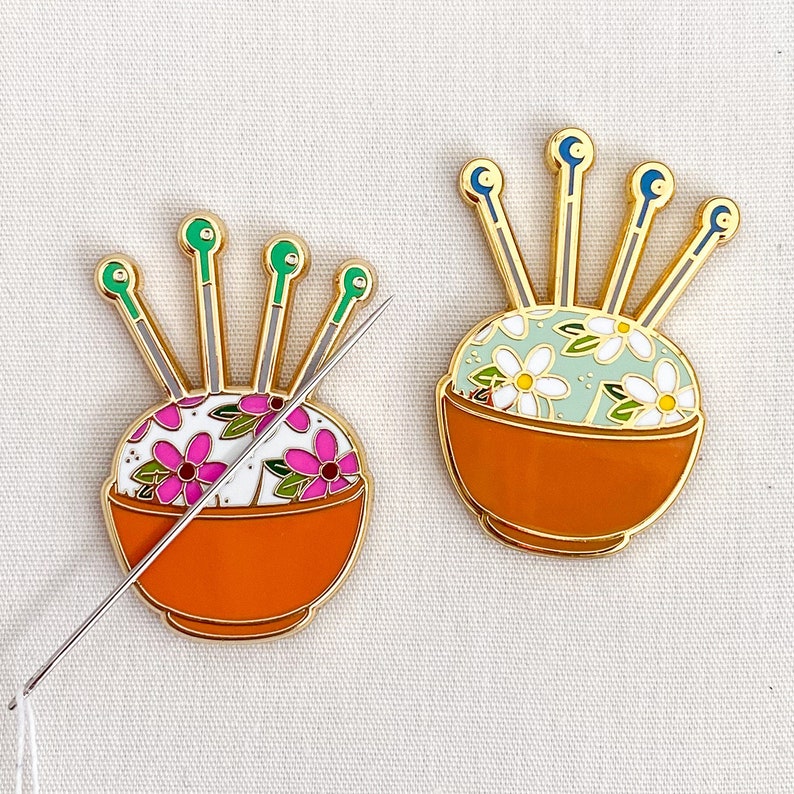 May include: Two gold pincushions with a floral design. Each pincushion has a bowl shape with a needle holder and a floral pattern. One pincushion has a pink and white floral pattern and the other has a green and white floral pattern.