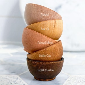 May include: A stack of five wooden bowls in various shades of brown, from light to dark. The bowls are labeled with their wood types: Natural, Teak Oil, Colonial Maple, Golden Oak, and English Chestnut.