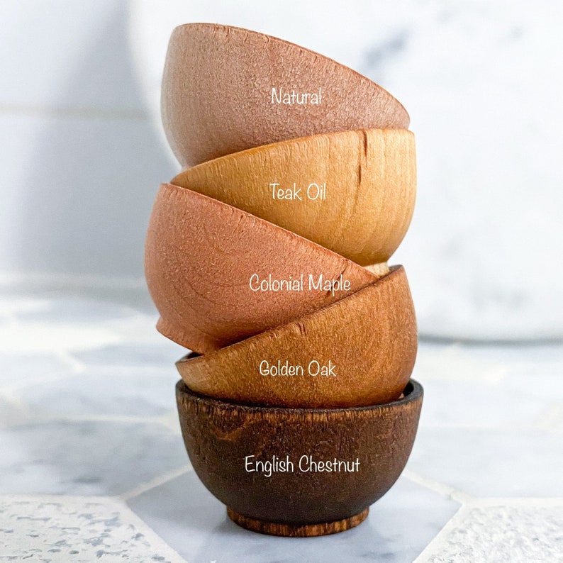 May include: A stack of five wooden bowls in various shades of brown, from light to dark. The bowls are labeled with their wood types: Natural, Teak Oil, Colonial Maple, Golden Oak, and English Chestnut.
