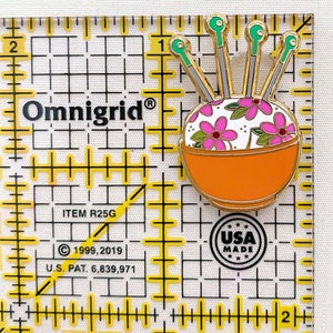 May include: A gold-coloured pin featuring a white bowl filled with colourful flowers and green and gold needles. The pin is on a white background with a yellow grid. The text "Omnigrid&reg;" is visible in the top left corner. The text "ITEM R25G" is visible in the bottom left corner. The text "&copy; 1999, 2019" is visible in the bottom left corner. The text "U.S. PAT. 6,839,971" is visible in the bottom left corner. The text "USA MADE" is visible in the bottom right corner.
