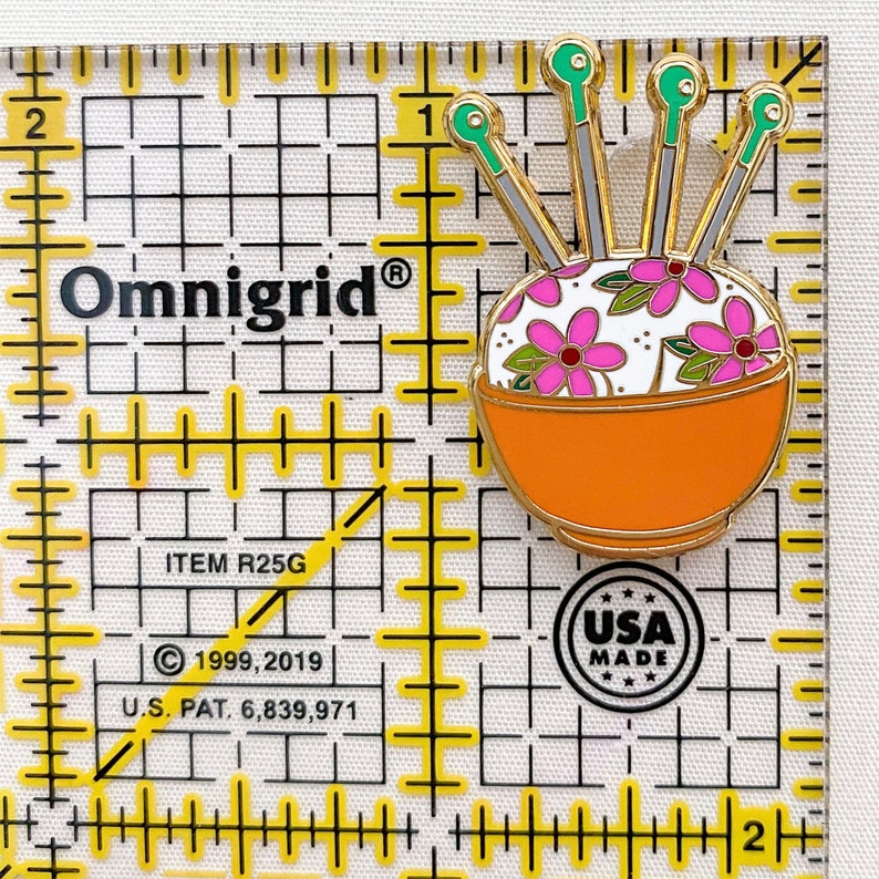 May include: A gold-coloured pin featuring a white bowl filled with colourful flowers and green and gold needles. The pin is on a white background with a yellow grid. The text "Omnigrid&reg;" is visible in the top left corner. The text "ITEM R25G" is visible in the bottom left corner. The text "&copy; 1999, 2019" is visible in the bottom left corner. The text "U.S. PAT. 6,839,971" is visible in the bottom left corner. The text "USA MADE" is visible in the bottom right corner.