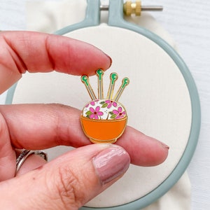 May include: A gold-tone enamel pin of a pincushion with pink flowers and green stems. The pincushion is in a small orange bowl.