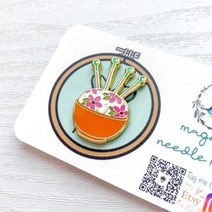 May include: A gold enamel pin of a needle minder with a white and orange bowl with pink and green flowers and five gold needles.