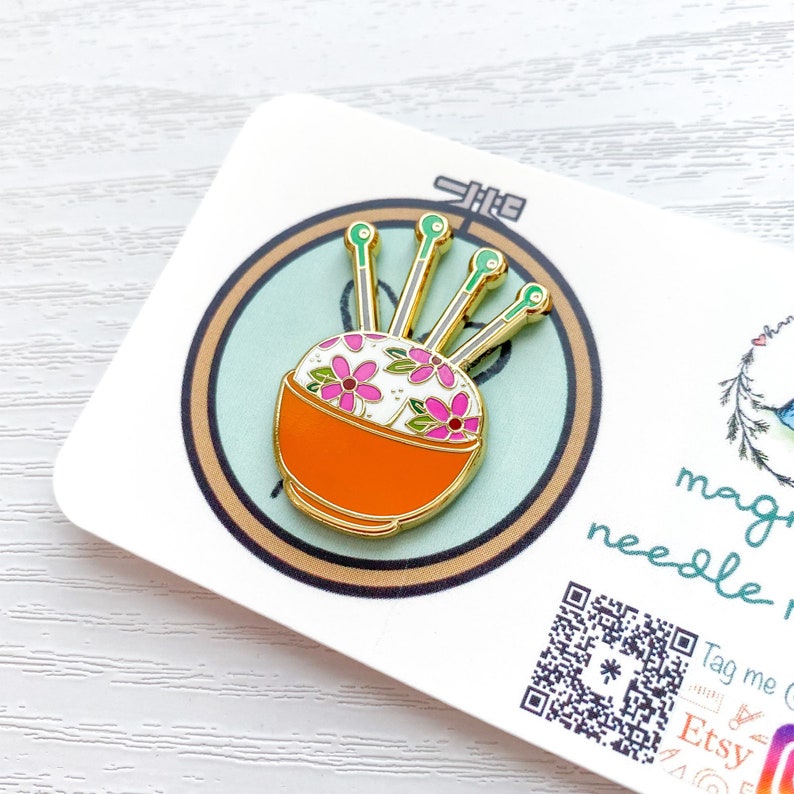 May include: A gold enamel pin of a needle minder with a white and orange bowl with pink and green flowers and five gold needles.