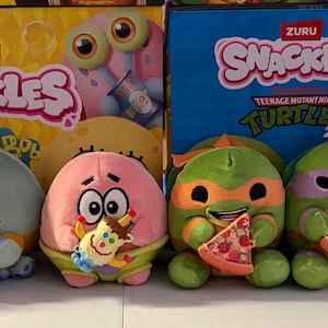 May include: A collection of plush toys featuring characters from SpongeBob SquarePants and Teenage Mutant Ninja Turtles. The toys are colorful and round, some holding miniature food items. Boxes with the "ZURU Snackles" logo are in the background.
