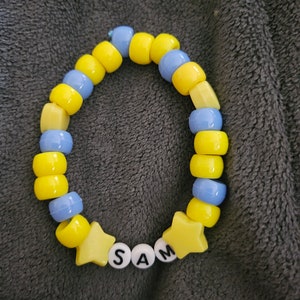 May include: A yellow and blue beaded bracelet with the name "SAM" spelled out in white letters on yellow star-shaped beads.