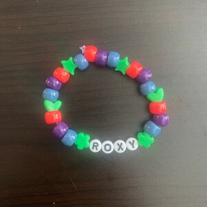 May include: A colorful beaded bracelet with green star and heart charms. The bracelet spells out the name "ROXY" in black letters.