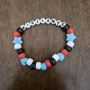 May include: A beaded bracelet with the word "VIGILANTE" spelled out in white letter beads. The bracelet features a pattern of red, black, white, and blue beads, including star-shaped and round beads. The bracelet is on a wooden surface.