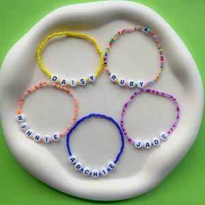 May include: Five colourful beaded bracelets with white letter beads spelling out the names "Daisy", "Ruby", "Winnie", "Archie", and "Jade".