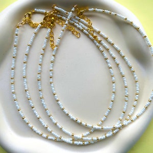 May include: Four white beaded necklaces with gold accents. The necklaces are layered on a white surface.
