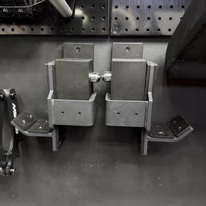 May include: A pair of black metal gym equipment attachments mounted to a wall panel. Each unit features a rectangular block, a protruding silver metal pin, and a lower angled support bracket with pre-drilled holes for secure installation.
