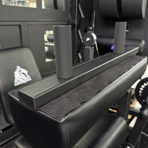 May include: A black gym equipment attachment featuring a rectangular base with two vertical, cylindrical handles. The handles have a textured, knurled grip surface. The base is covered in a black fabric material and is mounted onto a padded black seat.