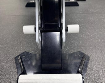May include: A black metal gym equipment component featuring a horizontal white textured handle at the base and two smaller white cylindrical knobs extending from the sides of the central vertical frame, all set against a dark gray floor surface.