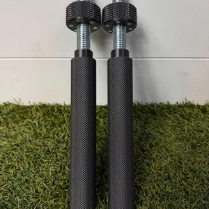 May include: Two identical cylindrical metal tools with a black knurled grip surface. Each tool features a silver threaded rod extending from the top, topped with a circular black knurled adjustment knob that has small silver circular accents.