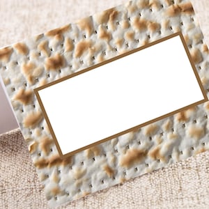 May include: A matzah card with a white rectangular label framed in brown. The matzah has a light beige color with darker brown spots and small perforations. The card rests on a textured, light beige surface.