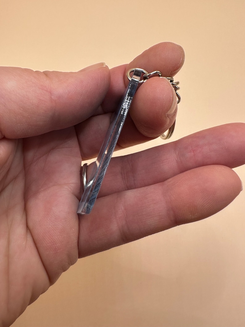 Pode incluir: Um chaveiro de acr&iacute;lico transparente com uma corrente de metal prateada. O chaveiro tem a forma de um ret&acirc;ngulo com cantos arredondados.