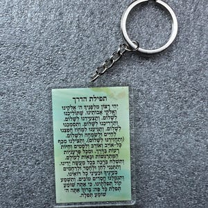 Pode incluir: Um chaveiro branco e verde com um chaveiro prateado. O chaveiro apresenta uma ora&ccedil;&atilde;o hebraica, "Tefilat HaDerech", que se traduz como "Ora&ccedil;&atilde;o para a Viagem".