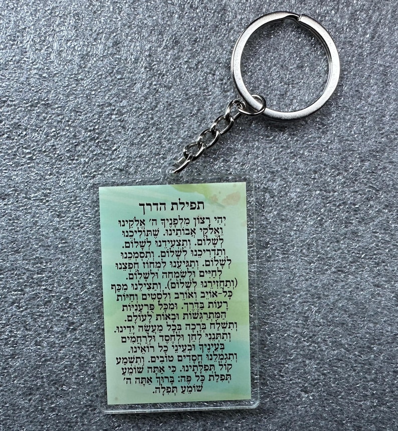 Pode incluir: Um chaveiro branco e verde com um chaveiro prateado. O chaveiro apresenta uma ora&ccedil;&atilde;o hebraica, "Tefilat HaDerech", que se traduz como "Ora&ccedil;&atilde;o para a Viagem".