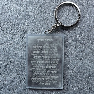 Pode incluir: Um chaveiro de acr&iacute;lico transparente com um anel de metal prateado. O chaveiro apresenta uma ora&ccedil;&atilde;o hebraica, "Tefilat HaDerech", que se traduz como "Ora&ccedil;&atilde;o do Caminho".