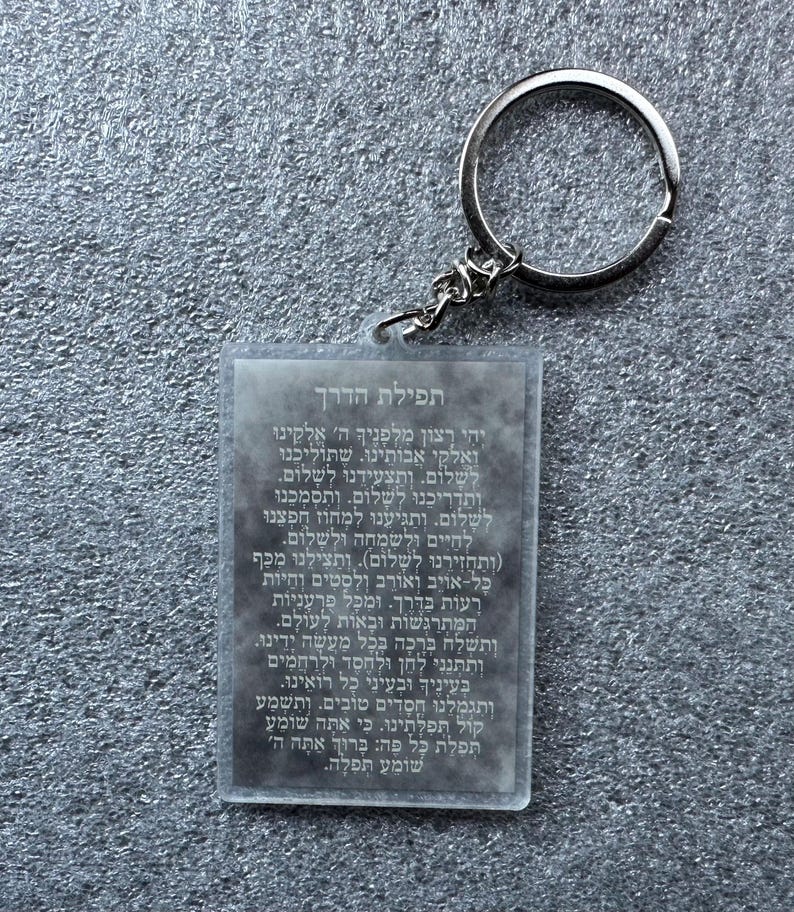 Pode incluir: Um chaveiro de acr&iacute;lico transparente com um anel de metal prateado. O chaveiro apresenta uma ora&ccedil;&atilde;o hebraica, "Tefilat HaDerech", que se traduz como "Ora&ccedil;&atilde;o do Caminho".