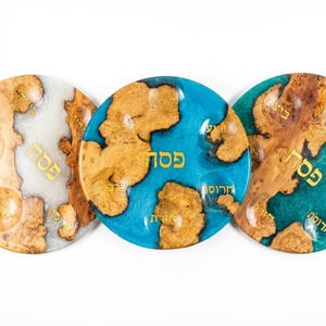 May include: Three round decorative plates with a wood and resin design. The plates feature a combination of wood grain patterns and colored resin, including white, blue, and teal. Gold Hebrew text is visible on each plate.