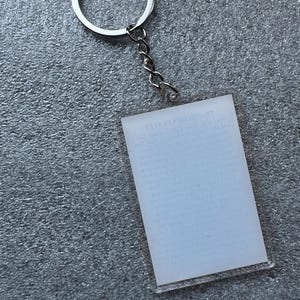 Pode incluir: Um chaveiro de acr&iacute;lico transparente com um anel e uma corrente de metal prateado. O chaveiro tem um ret&acirc;ngulo branco em branco com o texto "THE GREATEST GIFT" impresso nele.