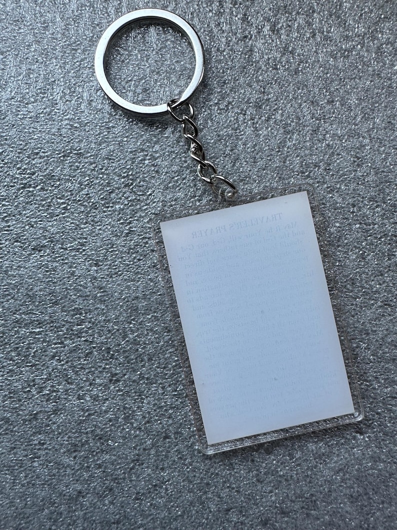 Pode incluir: Um chaveiro de acr&iacute;lico transparente com um anel e uma corrente de metal prateado. O chaveiro tem um ret&acirc;ngulo branco em branco com o texto "THE GREATEST GIFT" impresso nele.