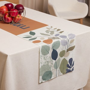 Puede incluir: Camino de mesa con un diseño botánico con hojas verdes, azules y naranjas. El camino de mesa tiene una sección rectangular con la palabra "KU" en azul. Una cesta de alambre dorada contiene manzanas rojas.