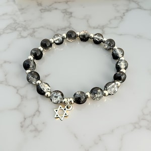 May include: A beaded bracelet featuring black and silver-toned beads. The bracelet has a silver-colored charm in the shape of a Star of David. The beads are strung together with silver-colored metal. The bracelet is displayed on a white and gray marble surface.