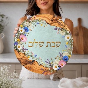 May include: A round wooden serving board with a light blue centre featuring the Hebrew text "Shabbat Shalom" in gold lettering, surrounded by a colourful floral border of daisies, cornflowers, and lavender on a natural wood grain edge.