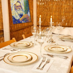 May include: A dining table set for a special occasion, featuring white plates with gold rims, silverware, and wine glasses. Two lit candles in silver holders add to the ambiance. A painting hangs on the bamboo wall.