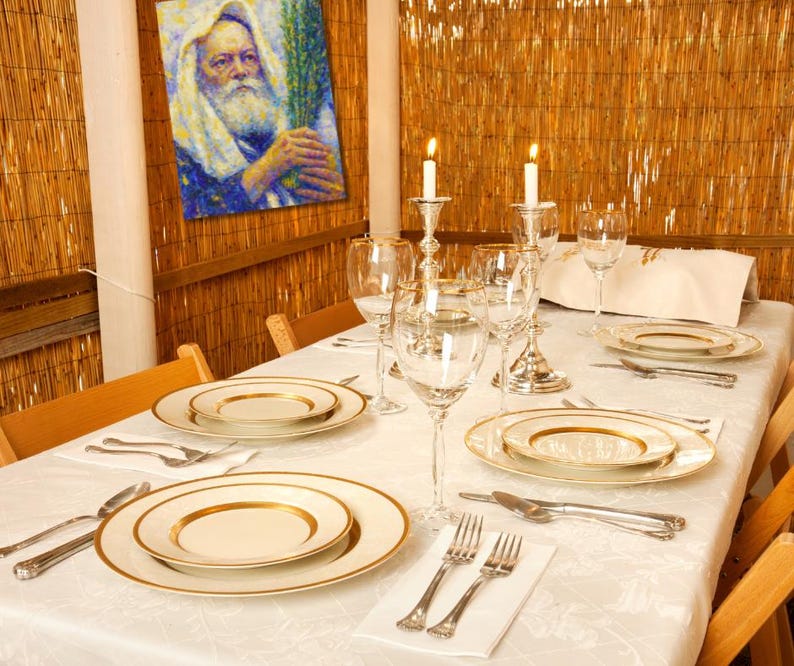 May include: A dining table set for a special occasion, featuring white plates with gold rims, silverware, and wine glasses. Two lit candles in silver holders add to the ambiance. A painting hangs on the bamboo wall.