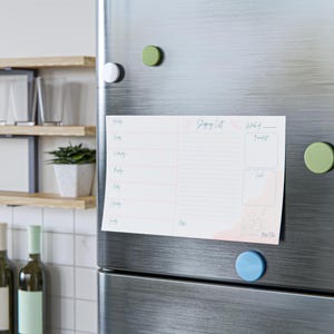 Puede incluir: Un bloc de notas magnético blanco para la lista de la compra con detalles en rosa e ilustraciones florales está adherido a un frigorífico de acero inoxidable con imanes de colores. El bloc de notas incluye secciones para cada día de la semana, una lista de la compra y planificación de comidas.