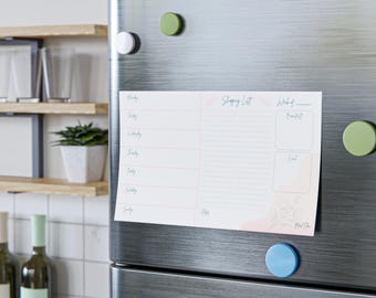 Op de afbeelding: Een witte magnetische boodschappenlijstnotitieblok met roze accenten en bloemenillustraties is met kleurrijke magneten aan een roestvrijstalen koelkast bevestigd. Het notitieblok bevat secties voor elke dag van de week, een boodschappenlijst en maaltijdplanning.