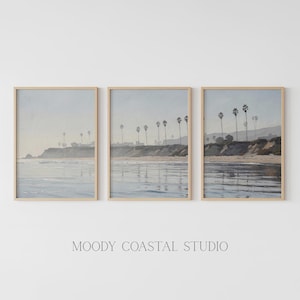 May include: Three framed coastal landscape prints. Each print features a beach scene with palm trees, ocean waves, and a hazy sky. The frames are a light wood color. The text "MOODY COASTAL STUDIO" is below the prints.
