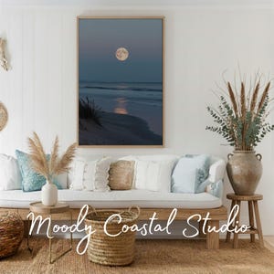 Könnte beinhalten: Ein gerahmter vertikaler Wandkunstdruck, der einen Vollmond über einem dunklen Meeresstrand in der Dämmerung zeigt, mit einer Spiegelung auf dem Wasser. Die Szene zeigt Sanddünen und ruhige Wellen in dunklen Blau- und Grautönen sowie goldenem Licht.