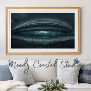 Puede incluir: Una impresión enmarcada de una dramática escena de tormenta costera. La imagen captura una oscura y arremolinada formación de nubes sobre el océano, con relámpagos que iluminan el agua. La impresión está en un marco de madera clara.