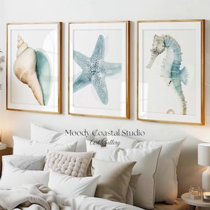 Puede incluir: Tres cuadros de pared enmarcados con ilustraciones en acuarela de una concha, una estrella de mar y un caballito de mar en tonos azul y beige. El texto Moody Coastal Studio Art Gallery está centrado bajo los cuadros sobre una cama.