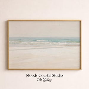 May include: A horizontal landscape painting in a thin gold frame depicting a serene beach scene with soft beige sand, gentle turquoise waves, and a pale sky. The text Moody Coastal Studio Art Gallery is printed in black below the framed artwork.