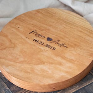 May include: A round wooden serving tray with a natural wood finish. The tray is engraved with the names "Megan & Austin" and the date "09.21.2019".