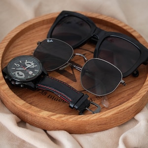 May include: A wooden tray with a black watch, a pair of black sunglasses, and a pair of silver sunglasses.