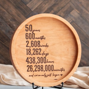 May include: A round wooden plaque with a black metal stand. The plaque has a message engraved on it that reads "50 years, 600 months, 2,608 weeks, 18,262 days, 438,300 hours, 26,298,000 minutes, and we've only just begun."