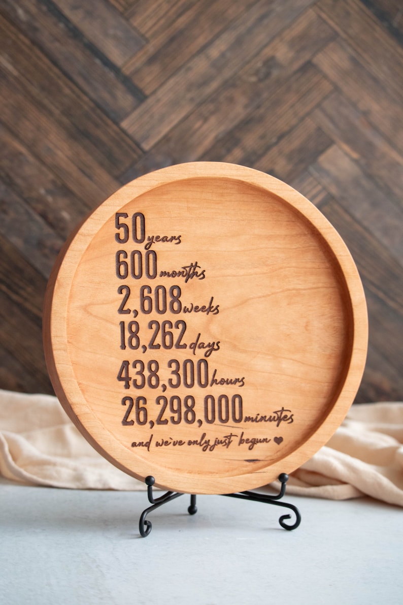 May include: A round wooden plaque with a black metal stand. The plaque has a message engraved on it that reads "50 years, 600 months, 2,608 weeks, 18,262 days, 438,300 hours, 26,298,000 minutes, and we've only just begun."