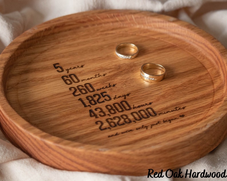 May include: A wooden ring dish with the inscription "5 years, 60 months, 260 weeks, 1,825 days, 43,800 hours, 2,628,000 minutes, and our story just begun." Two gold rings are placed on the dish.