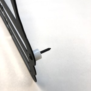 May include: Close-up of a black metal wall decor with a decorative design. A black screw is inserted into a white plastic spacer, ready for mounting. The decor has a cut-out design.