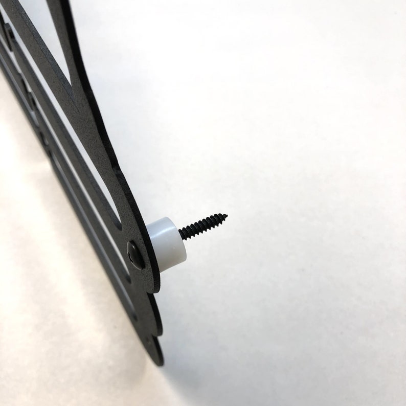 May include: Close-up of a black metal wall decor with a decorative design. A black screw is inserted into a white plastic spacer, ready for mounting. The decor has a cut-out design.