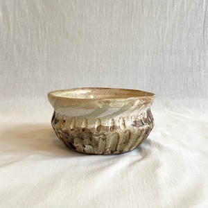 Puede incluir: Un cuenco de cerámica blanca con esmalte marrón y verde. El cuenco tiene un borde texturizado y una forma ligeramente redondeada.
