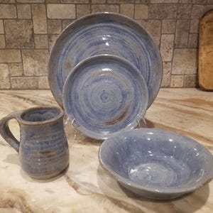 Puede incluir: Un juego de vajilla de cerámica azul hecho a mano. El juego incluye un plato grande, un plato más pequeño, un cuenco y una taza. Las piezas tienen un aspecto rústico y texturizado con patrones en espiral visibles.